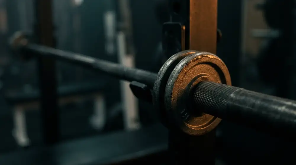 Primer plano de dos discos pequeños de hierro fraccionado montados en la manga moleteada de una barra olímpica, con el gimnasio a oscuras desenfocado detrás.